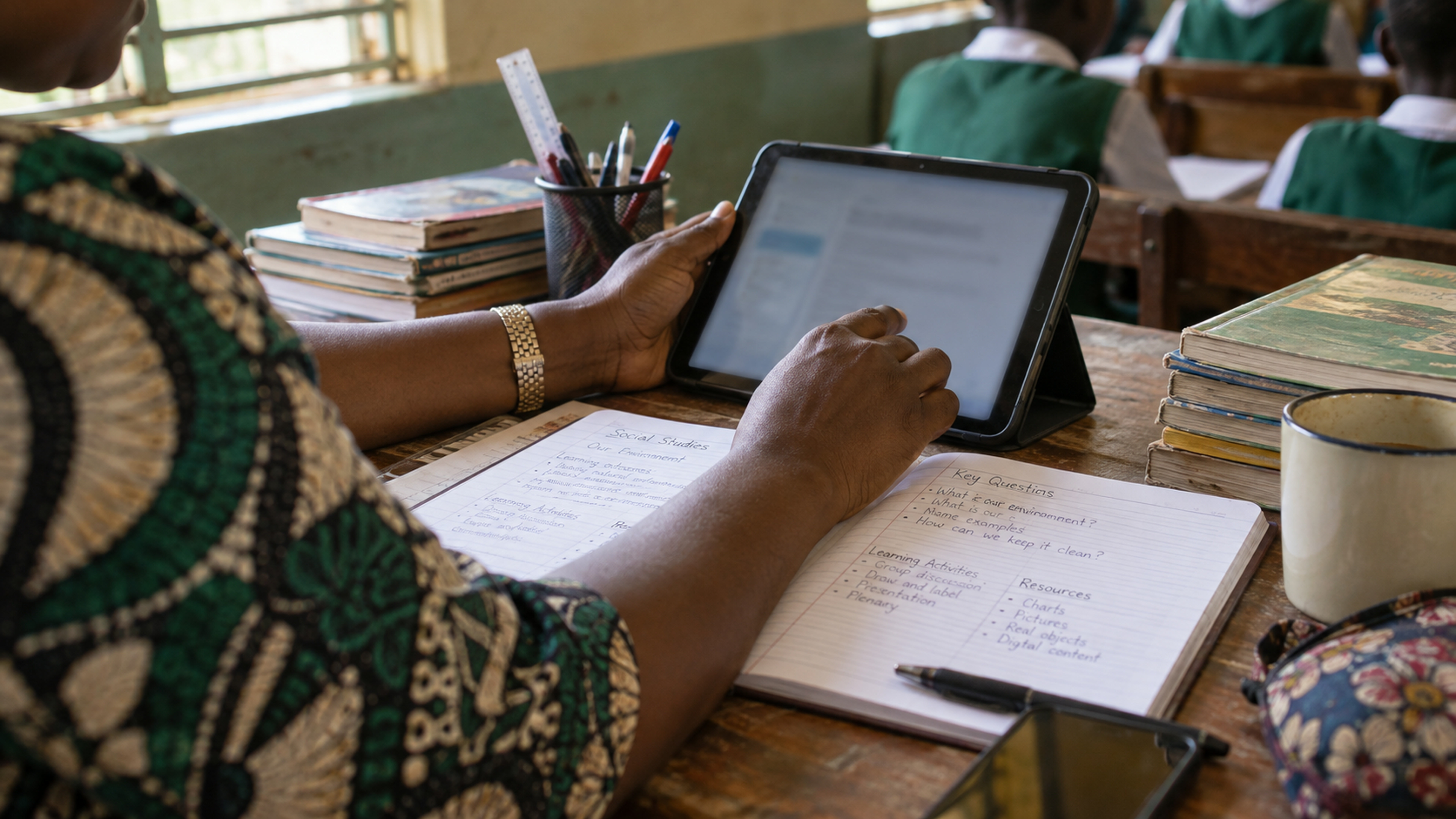 A teacher’s hands on a tablet beside handwritten lesson notes.
