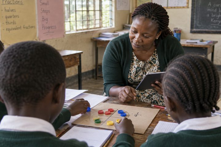 A teacher glances at a tablet while learners work in a small group.