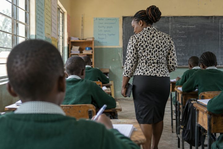 A teacher walking between desks during a lesson, seen from behind the nearest child.